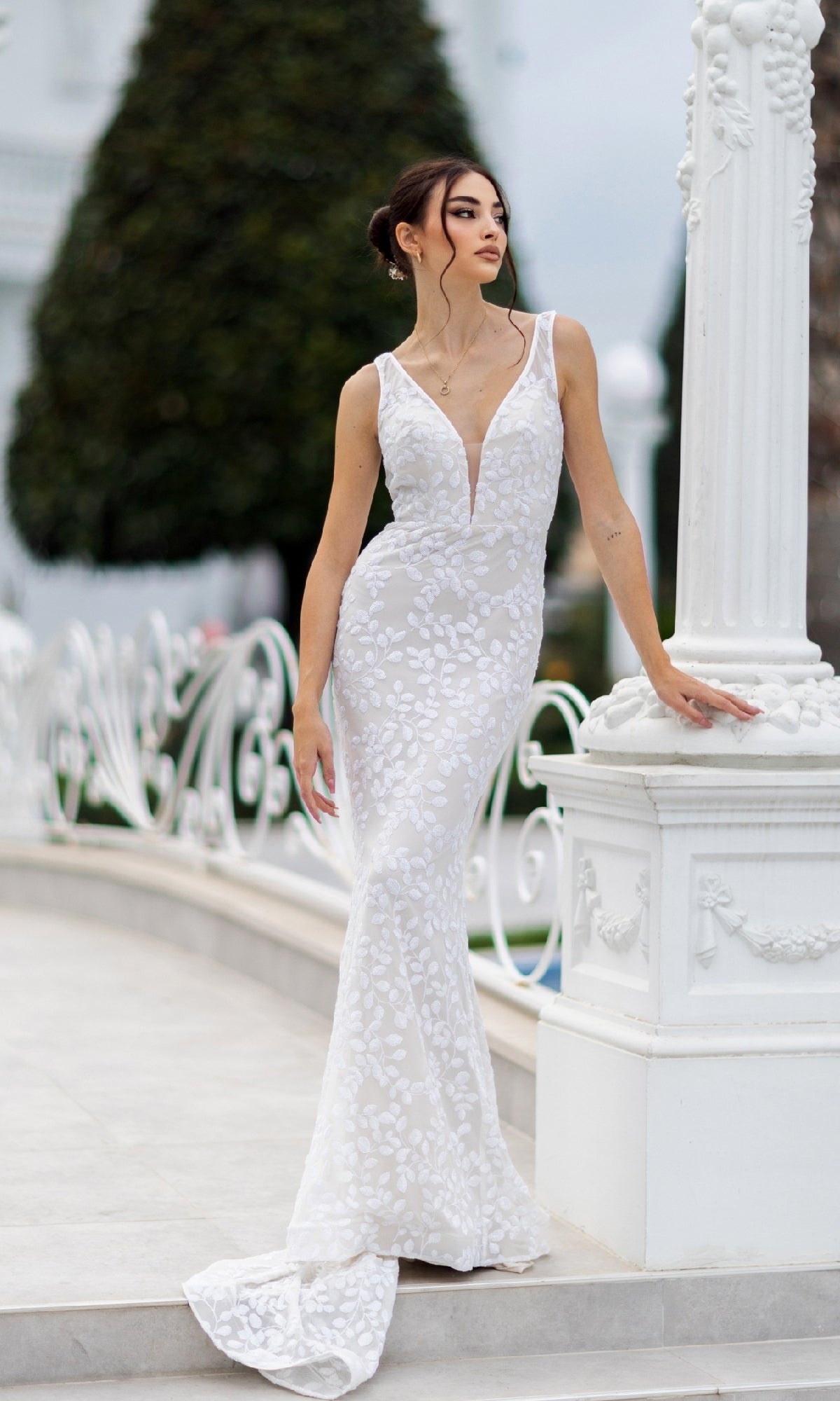 A woman wearing the Tina Holly TW108 long white sequin formal dress with a fitted, sleeveless mermaid silhouette and deep V-neckline stands by a white column outdoors.